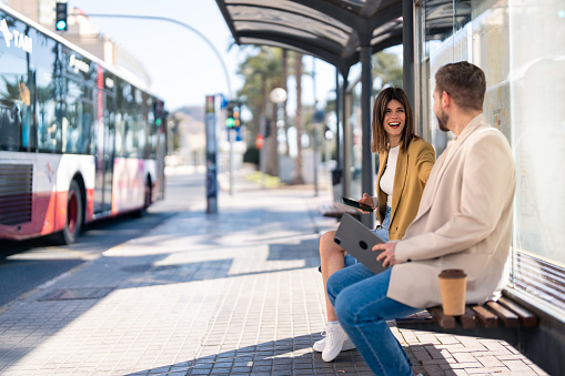 Two cheerful friends young woman and man sitting at bus station while city bus is passing by on the street, having a conversation, laughing, holding mobile phone and digital tablet. Photo with copy space.