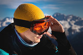 Portrait of a professional skier athlete in a blue jacket, black and orange mask looks to the side. Background with snow covered mountains of ski resort