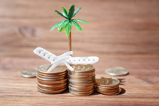 Offshore tax haven concept: Toy beach lounger placed atop coins stacks under a palm tree figurine arranged on a wooden background. Business, tax planning and wealth management related backdrop.