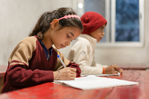 Indian children of elementary school age studying in the classroom, young girl wearing school uniform and writing on a notepad, learning and education concept, India.