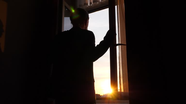 Man stands and looks out, then closes window sashes and drapes curtains