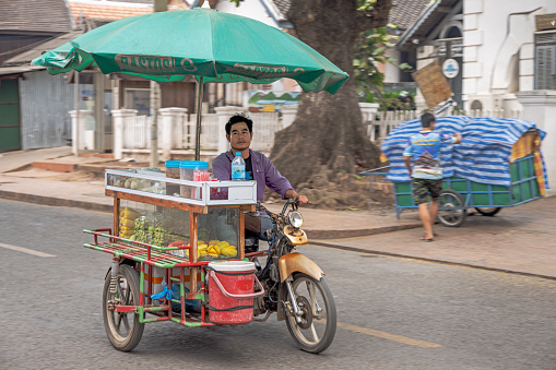 Luang Prabang, Laos - March 12th 2023: Street vendor with a shop on a small three wheeled motorcycle under a large umbrella