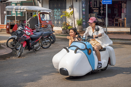 Luang Prabang, Laos - March 12th 2023: Two young female tourists on a funny motorcycle with sidecar