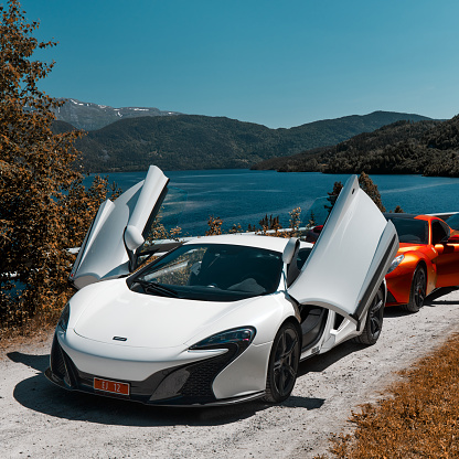 Road trip with supercars. Mclaren 650s, Ferrari F12  parked with open door on a gravel road by the lake. Gransherad, Norway. 04.06.2016