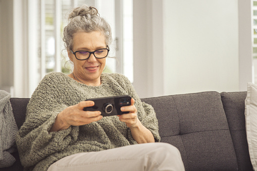 A beautiful woman is sitting comfortably on a sofa, at home playing games on her phone.