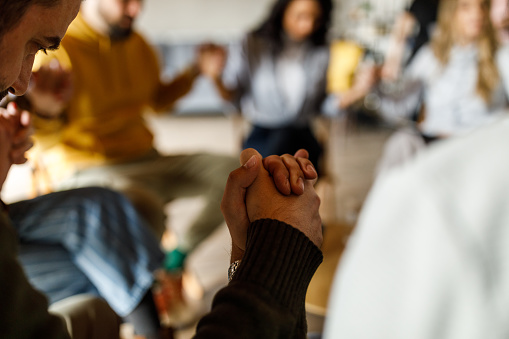 Close up shot of diverse group of adult people sitting in a circle at the group therapy session and holding hands.