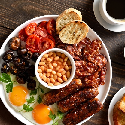 Stock photo showing close-up, elevated view of a white plate containing a fried breakfast with ramekin of baked beans, two sunny-side-up fried eggs, bacon rashers, sausages, grilled tomatoes, mushrooms and wholemeal toast.