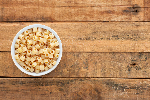 Top view of white bowl full of uncooked pasta shapes as square cubes over wooden table