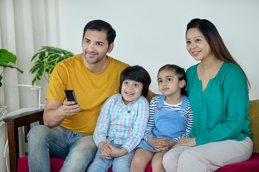 Stock image of happy Indian family sitting together at home - wearing casual clothes and smiling and laughing while watching a comedy show