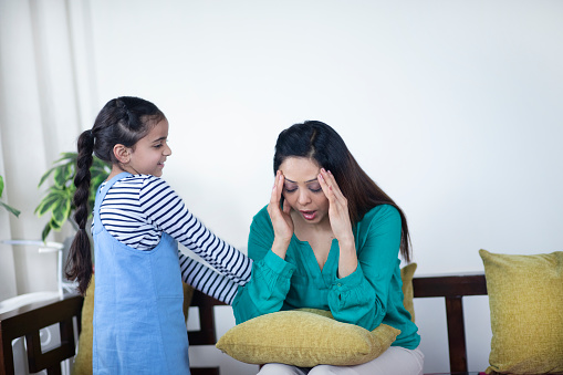 Smiling daughter holding young mother hand and asking her to play - stressed mother having headache, wearing casuals