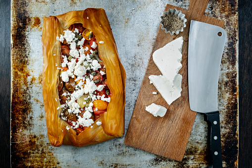 Crumbled feta cheese served with roasted tomatoes with filo pastry. Colour, horizontal format with some copy space.
Take 8 sheets of filo pastry, brushing with melted butter in-between each layer, on top of the last layer place some sliced cherry tomatoes, season with salt and pepper and add the capers, leave enough room around the edge to fold the edges over to make a “parcel”. Bake for 20 minutes until the filo pastry turns a rich golden brown colour, allow to cool on a wire tray then cut serve with crumbled pieces of feta cheese. Delicious!