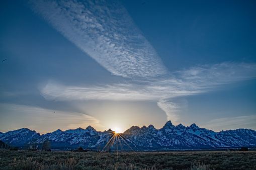 Teton mountain range with old barn and star burst sunlight and cloud stream in Yellowstone Ecosystem of western USA and North America.
