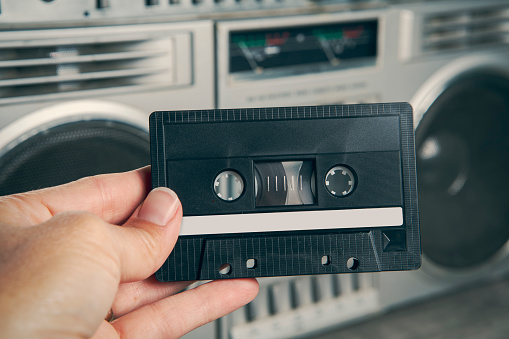 A woman's hand holding a cassette with a blank label with a boom box stereo blurred in the background.