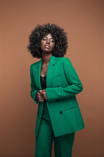 Portrait of fashionable young woman with afro hairstyle wearing green suit, looking away. Brown background.