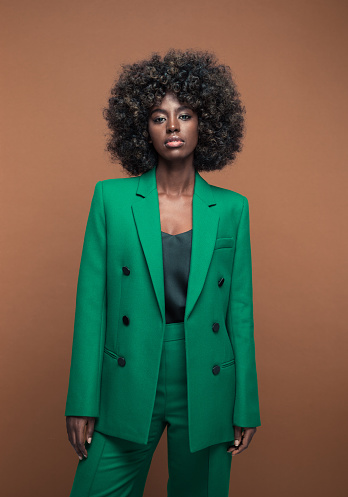 Portrait of fashionable young woman with afro hairstyle wearing green suit, looking at camera. Brown background.