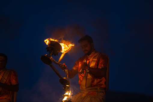 Varanasi, Uttar Pradesh, India - November 2022: Ganga aarti, Portrait of an young priest performing river ganges early morning aarti at assi ghat in traditional dress with fire flame traditionally.