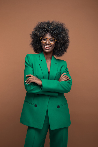 Portrait of fashionable young woman with afro hairstyle wearing green suit and brown glasses, standing with arms crossed and smiling at camera. Brown background.