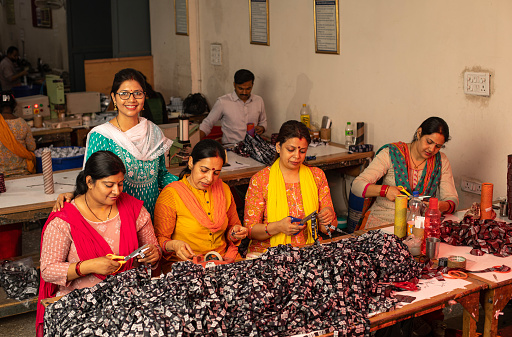Smiling female manager inspecting and guiding workers cutting printed garments on table at textile factory and representing women empowerment