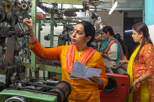 Mature woman with paper inspecting manufacturing equipment and female workers working at production line in textile factory and representing women empowerment