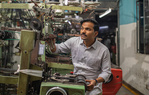 Serious mid adult Indian male mechanic dressed in casuals examining manufacturing equipment at textile factory