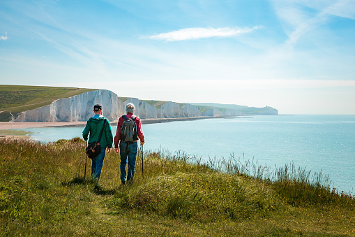 Rear view image depicting two men hiking together with the stunning backdrop of Seven Sisters cliffs in the south of England. It is a sunny day with a dazzling blue sky.