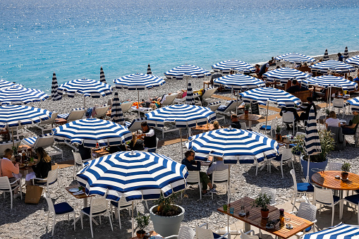 Nice, France - April 21, 2023: On a sunny day, white-blue umbrellas were spread on the beach on the Mediterranean Sea. There are people who enjoy the seaside charms in the shade of umbrellas