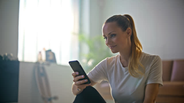 SLO MO Smiling young woman with ponytail sitting and texting with smart phone on living room floor at home. Lifestyle,domestic life,simple living. Shot in 8K Resolution.