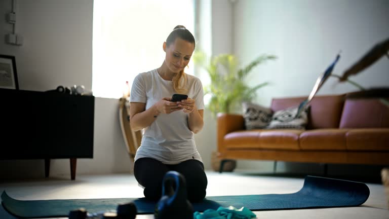 SLOW MOTION Fit young woman with dumbbells and kettlebell taking a break from home workout on yoga mat,texting with smart phone in living room. Healthy lifestyle,exercise,fitness,domestic life. Shot in 8K Resolution.