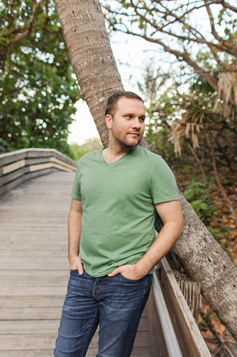 A 31-Year-Old American Man with Short Brown Hair & Green Eyes Dressed in Classic Green V-Neck T-Shirt & Navy Denim Jeans, Enjoying Nature on a Boardwalk in a Woodsy Forest in South Florida