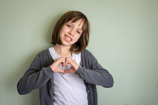 Child girl showing heart shape whit her hands while standing against wall and looking at camera indoors