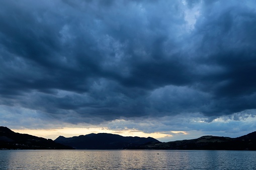 An expansive body of water with majestic mountain peaks in the backdrop, shrouded in a cloudy sky
