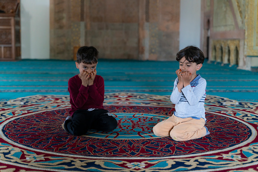 brothers sitting in the mosque praying with open hands. 
There is no one in the mosque except the child. Shot indoors in natural light with a full frame camera.