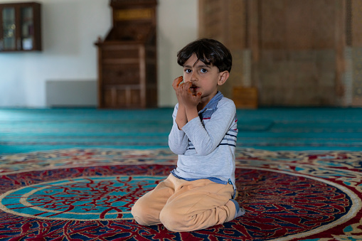 boy sitting in the mosque praying with open hands. 
There is no one in the mosque except the child. Shot indoors in natural light with a full frame camera.