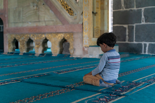 boy sitting in the mosque praying with open hands. \nThere is no one in the mosque except the child. Shot indoors in natural light with a full frame camera.