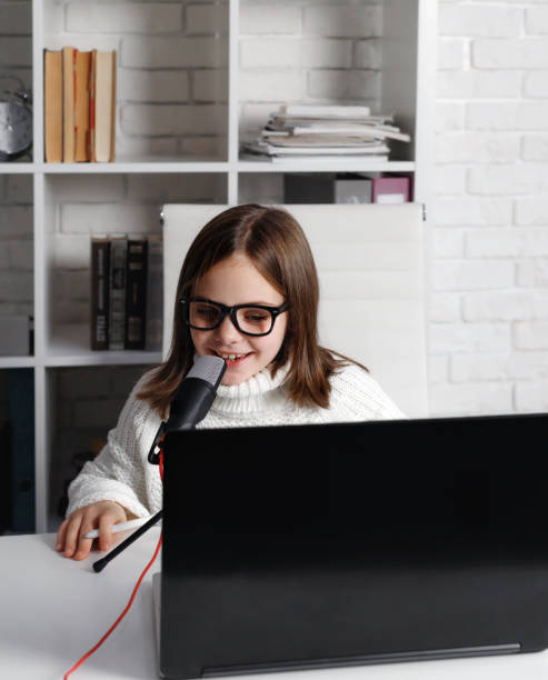 bambina in occhiali da vista che sorride e parla nel microfono, bambino che usa il computer portatile e guarda lo schermo durante la registrazione di audio o video - home schooling audio foto e immagini stock