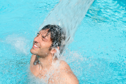 Happy young man having fun and relax under a water jet at swimming pool.