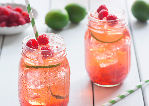 Raspberry lime iced tea in glasses with straws. Ingredients for making tea - including fresh limes and berries in a bowl - are on the white picnic table in the background.