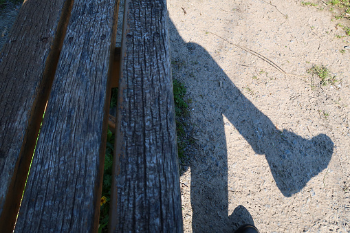 Dark Shadow Of A Woman's Legs, Sitting On A Wooden Park Bench On Dry Ground Outdoors In The Sun
