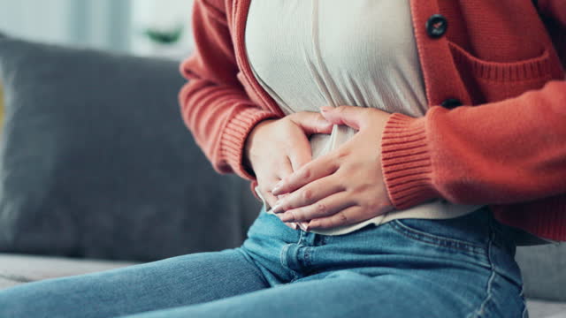 Stomach pain, woman hands and living room sofa with a female feeling abdomen cramps. Constipation, menstruation or ibs ache of a sick and suffering person holding lower belly on a home lounge couch