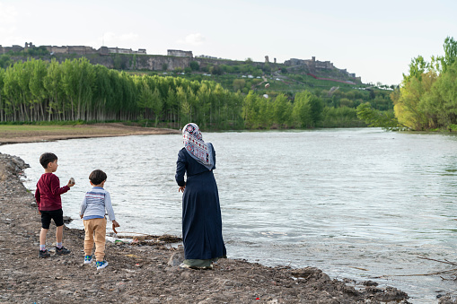 Siblings playing by the Tigris river in the city landscape of Diyarbakir surrounded by historical walls and their mother next to them. Tigris river flows through Hevsel gardens. the river in the foreground is the city on the hill in the background. Shot with a full-frame camera in daylight.