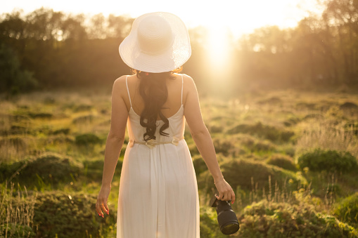 Photographer young woman walking in the forest at sunset. white dress and hat