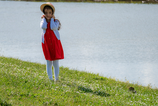 portrait of little girl in red dress with hat standing by the stream. In spring, the meadows are green with fresh grass.  Shot with a full-frame camera during the day.