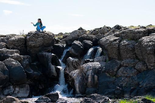 little girl sitting on the ground watching the view of the small waterfall flowing from the cliffs. In the spring season, water pours from the rocks.  Shot with a full-frame camera in daylight.