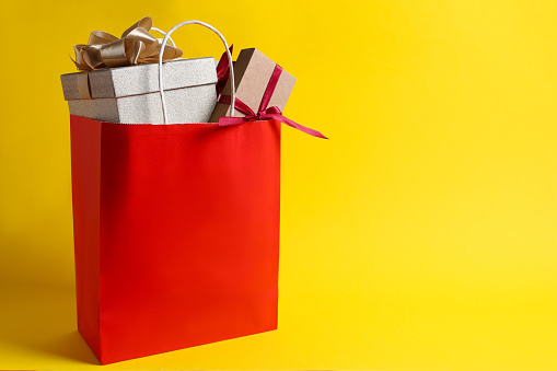 Red paper shopping bag full of gift boxes on yellow background. Space for text
