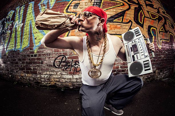 Hardcore Gangsta Rapper A stereotypical hip hop / gangsta / rapper character with baggy pants, gold chains (bling), and doo rag kneels in front of a graffiti covered brick wall, holding a large stereo cassette player (boom box) on his shoulder.  In his other hand is a 40 oz. bottle of malt liquor beer that he takes a big drink of.  Fish eye wide angle lens.  Horizontal with copy space. do rag stock pictures, royalty-free photos & images