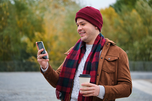 Latin young man with a coffee using a smart phone in the street in autumn.