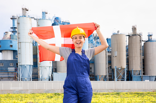 Cheerful female worker in hardhat with danish flag standing in front of factory
