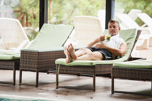 Full length shot of happy mature man in swimsuit, relaxing on a lounge chair at the poolside in a luxury spa resort and enjoying a refreshing beverage on his weekend getaway.