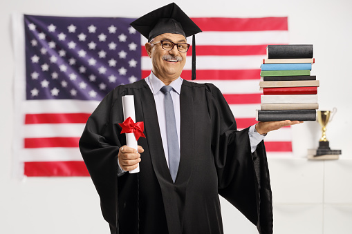 Mature man in a graduation gown holding a diploma and a pile of books in front of a usa flag