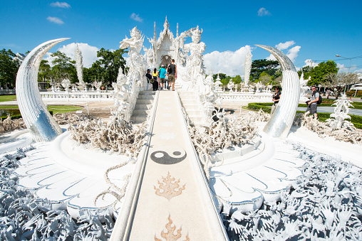 In Chiang Rai, Thailand visitors walk up the bridge designed with spiritual symbols including the Yin Yang towards the White Temple on a bright sunny day.
